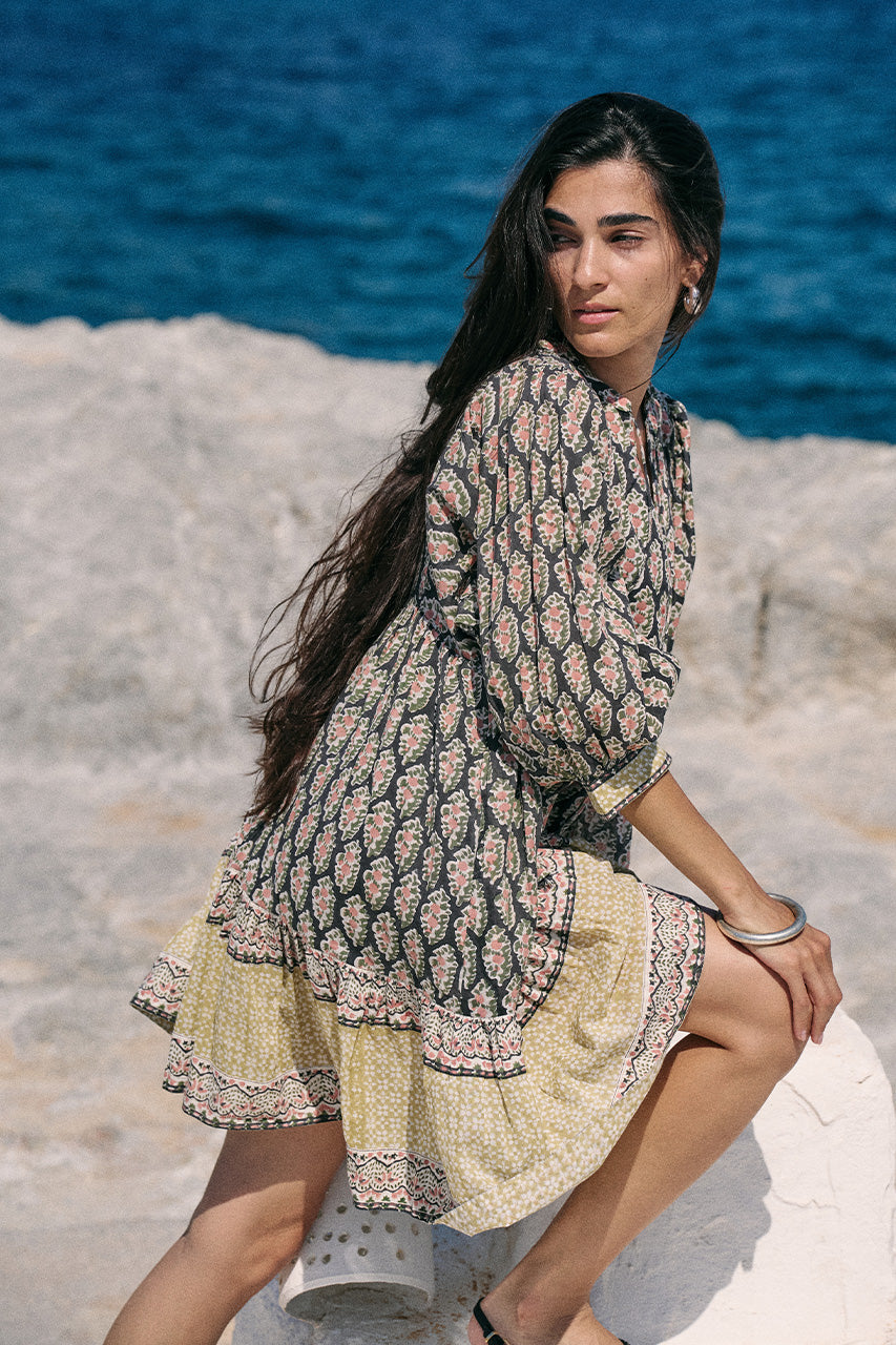 Woman posing on white coastal rock with ocean behind wearing the Daughters of India Bhoomi Mini Dress in Onyx, showing the tiered skirt hem detail with border print