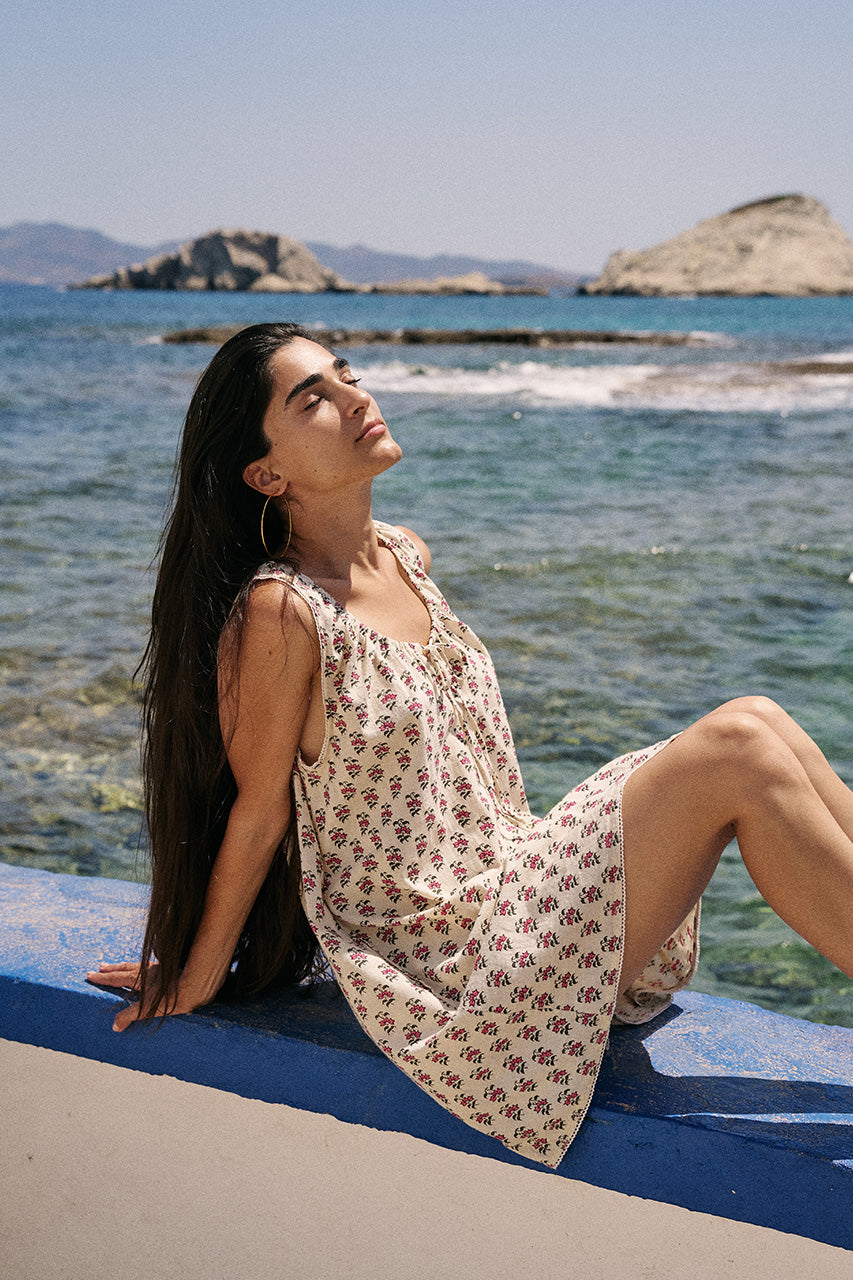 Sundress Mini Peony — reclining on blue seaside ledge basking in sun, hand block print peony floral cotton mini dress with ocean and rocky island behind | Daughters of India